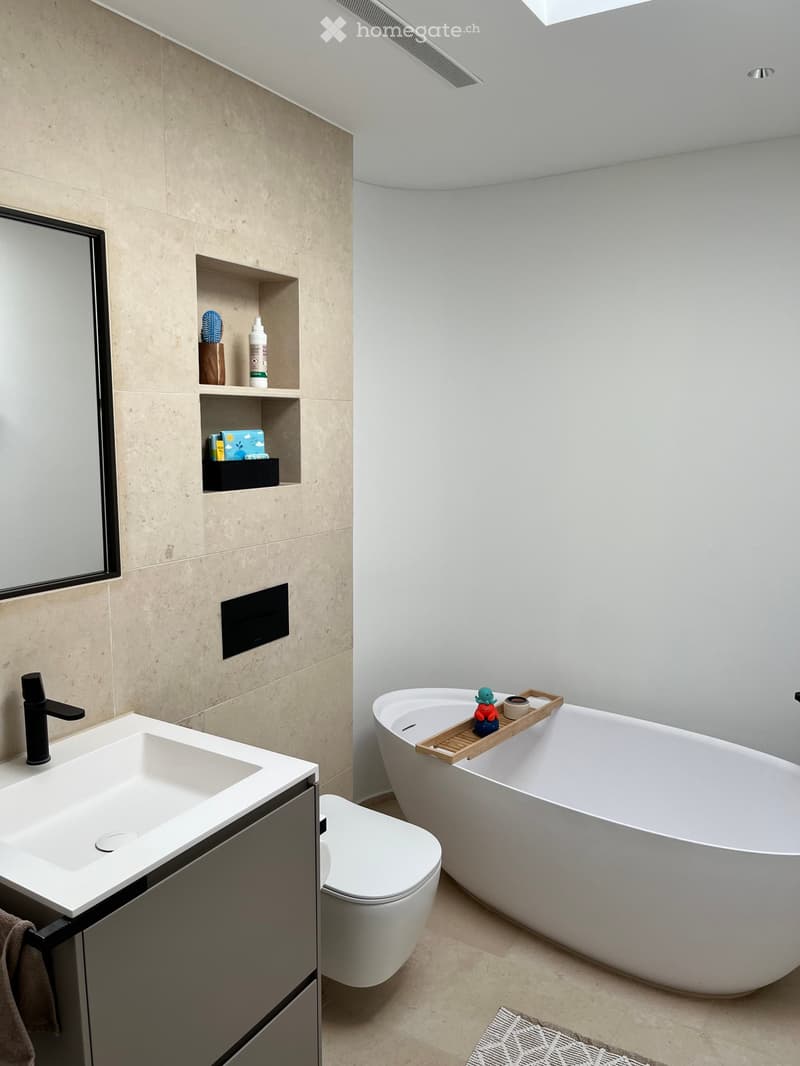 Modern bathroom with a large freestanding bathtub, a sleek sink with a modern faucet, a toilet, and a built-in shelf holding toiletries and a small decorative tray with a toy.