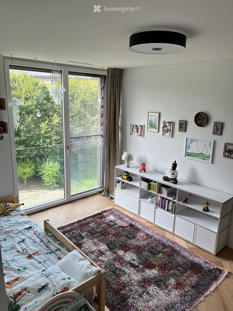 Bright bedroom with wooden floor, large window with view, bed with colorful bedding, shelf with books and toys, decorative items, and framed pictures on the wall.