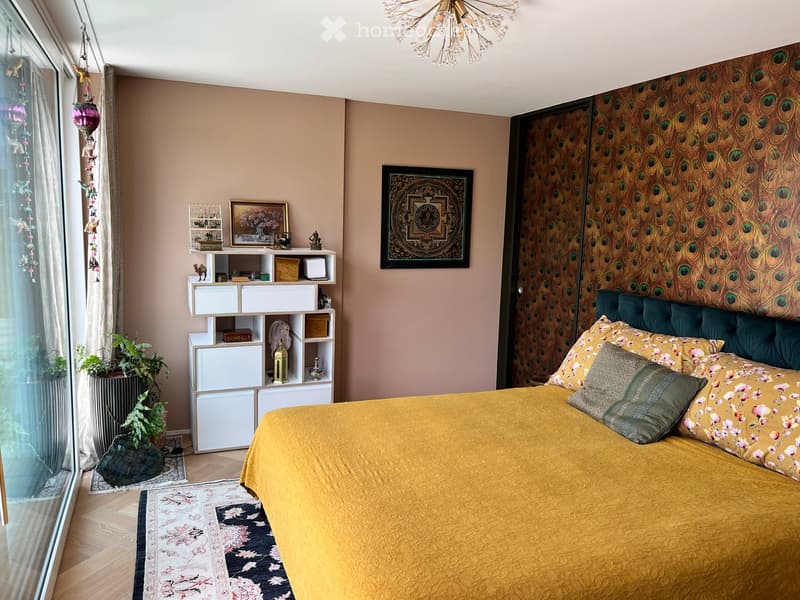 Cozy bedroom with a large bed covered in a mustard yellow duvet, teal headboard, and patterned pillows. A white shelving unit holds decorative items and books. The room features a patterned rug, framed artwork, and a chandelier hanging from the ceiling.