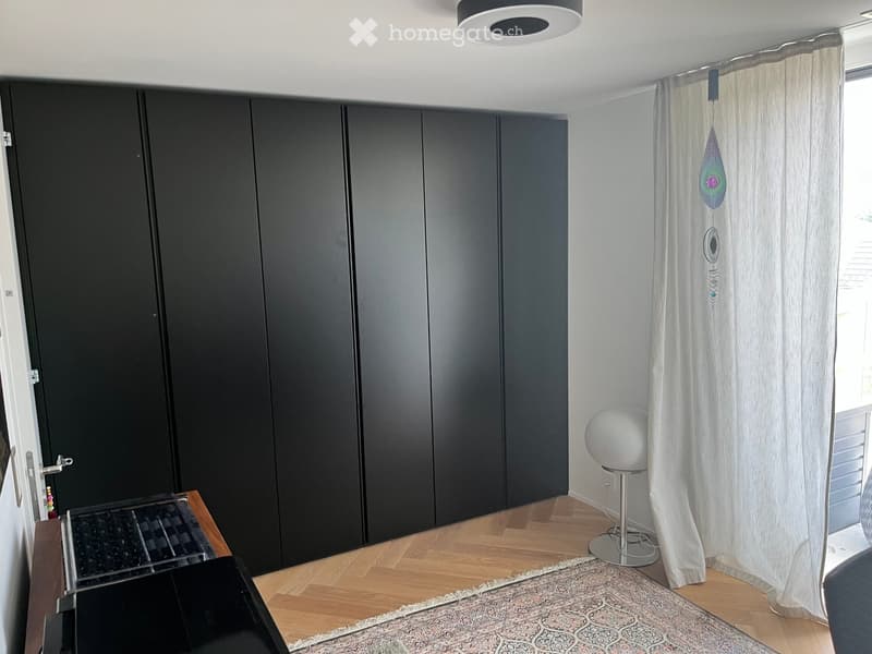 Modern room with wooden flooring, large black wardrobe, white stool, and decorative curtain with a hanging ornament.