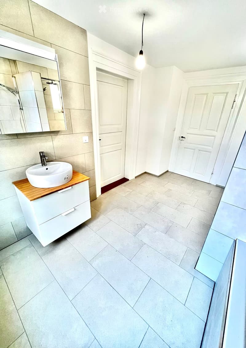 Modern bathroom with a white sink on a wooden countertop, a large mirror, and tiled walls and floor. Two white doors are visible, one leading to another room.