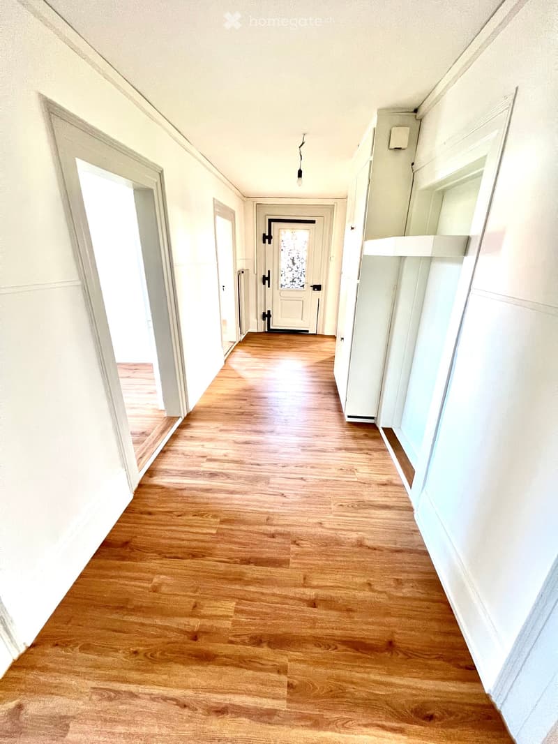 A long, narrow hallway with light wooden flooring and white walls. Several doors are visible along the sides, and the ceiling has a single hanging light fixture.