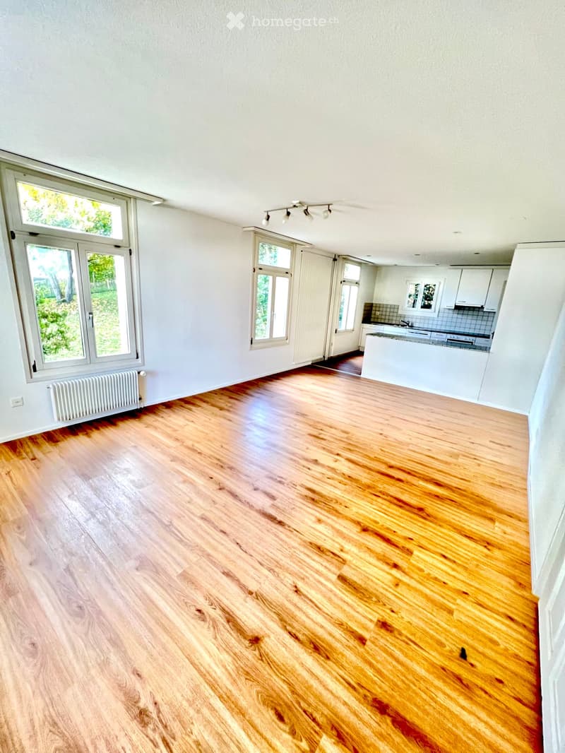 Empty room with wooden flooring, white walls, large windows, and a radiator. The adjacent kitchen is visible with white cabinets and appliances.