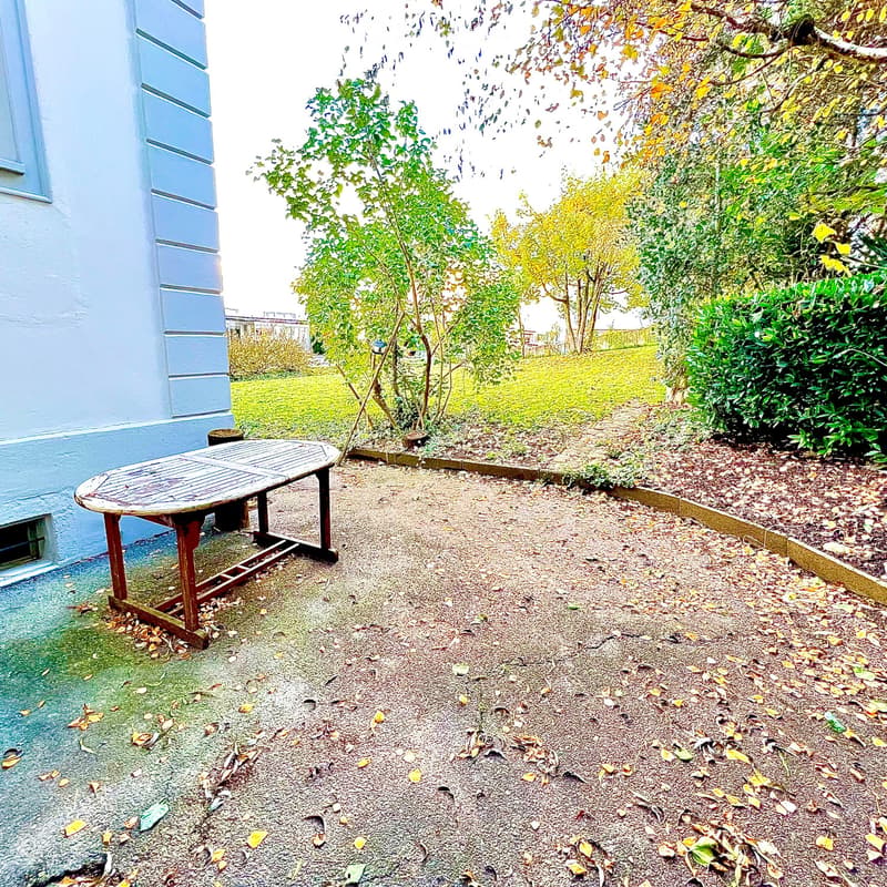 Garden area with gravel ground, a wooden table, and surrounding greenery including bushes and trees.