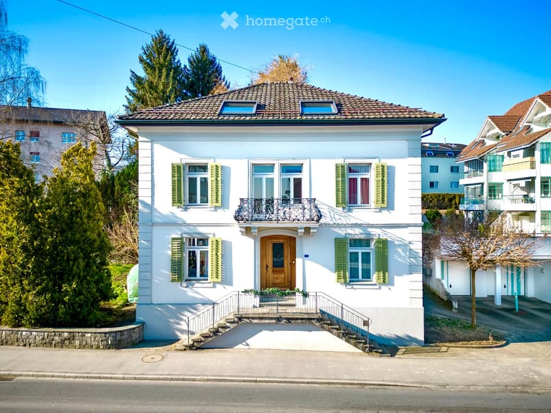 Two-story white house with green shutters, a balcony, and a wooden front door. The house has a tiled roof and is surrounded by trees and neighboring houses.