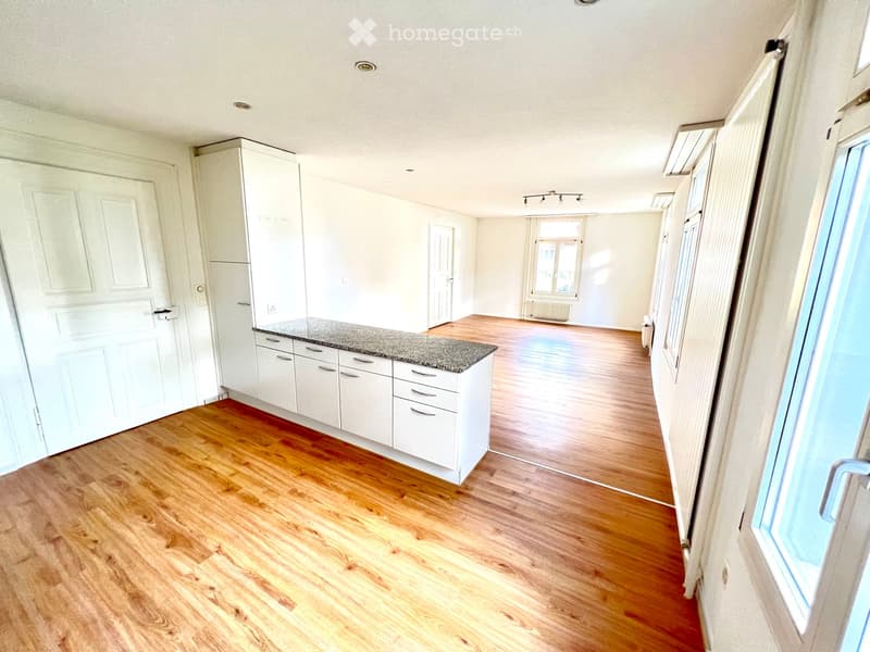 Empty kitchen with white cabinets, granite countertop, wooden flooring, and large windows allowing natural light.
