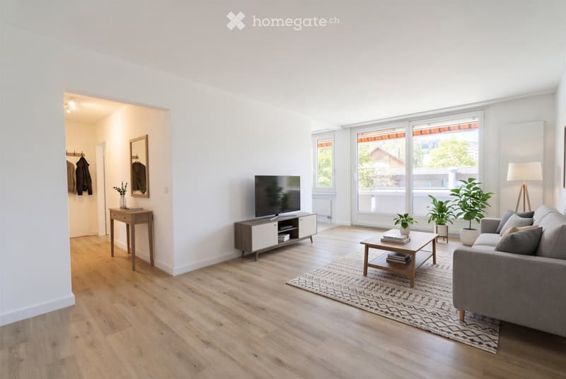 Bright living room with wooden flooring, large windows, grey sofa, coffee table, TV stand, side table, floor lamp, potted plants, and a coat rack in the adjacent hallway.