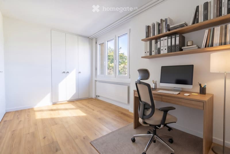A modern, well-lit study or home office with a wooden floor, large windows, a sleek desk setup, ergonomic chair, and built-in shelving holding books and binders.