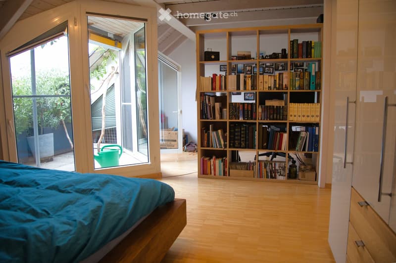 Bright bedroom with wooden floors, large windows, a bed with blue bedding, a bookshelf filled with books, and a chair.