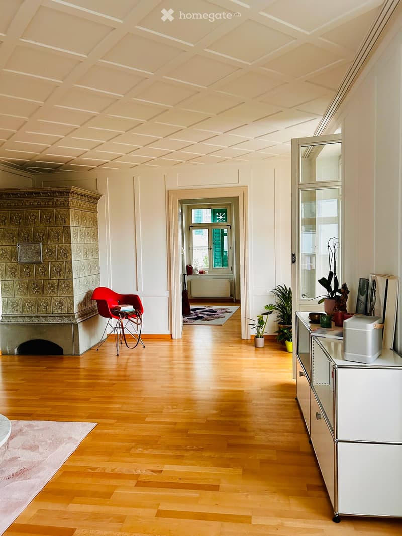 A spacious, well-lit room with wooden flooring, a fireplace, a red chair, a small table, a mirror, and several potted plants.