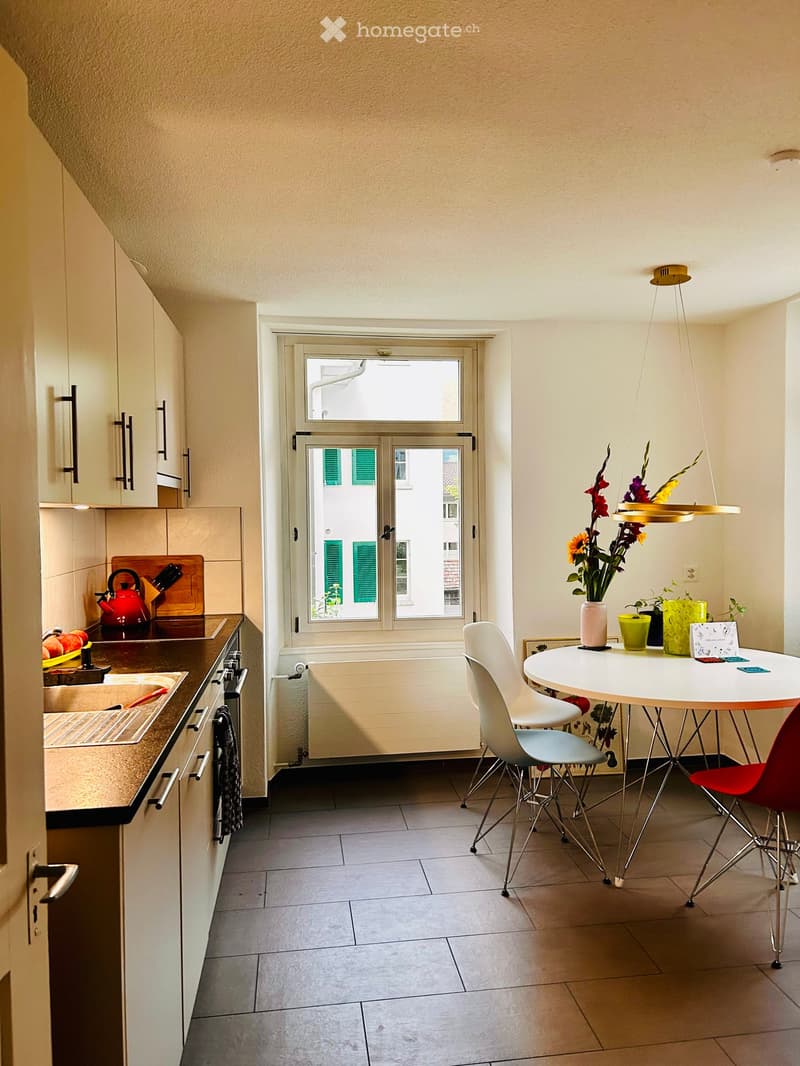 A modern, compact kitchen and dining area with white cabinets, a round dining table with chairs, a vase with flowers, and a small kitchen counter with a teapot and cutting board.