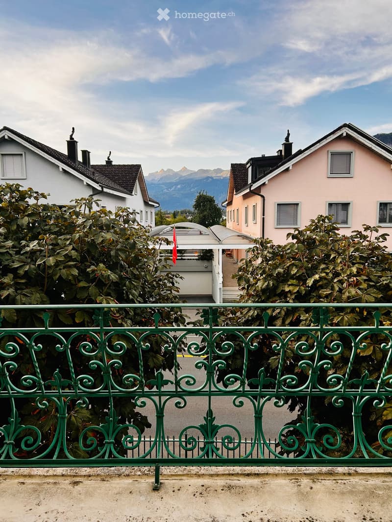 Two-story residential buildings with pitched roofs, surrounded by greenery and a scenic mountain view beyond.