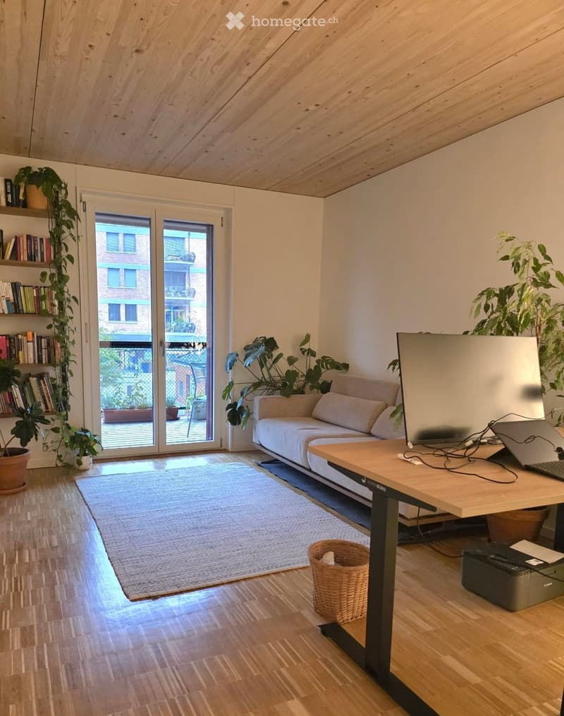 Bright living room with wooden floors, a white sofa, a wooden table with a laptop, a bookshelf, and several potted plants. A large sliding glass door leads to a balcony.