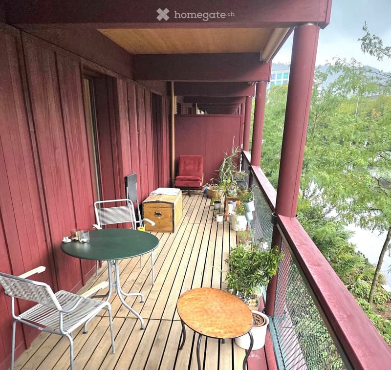 Wooden balcony with red wooden railings, featuring a green table, metal chairs, potted plants, and a small sofa.