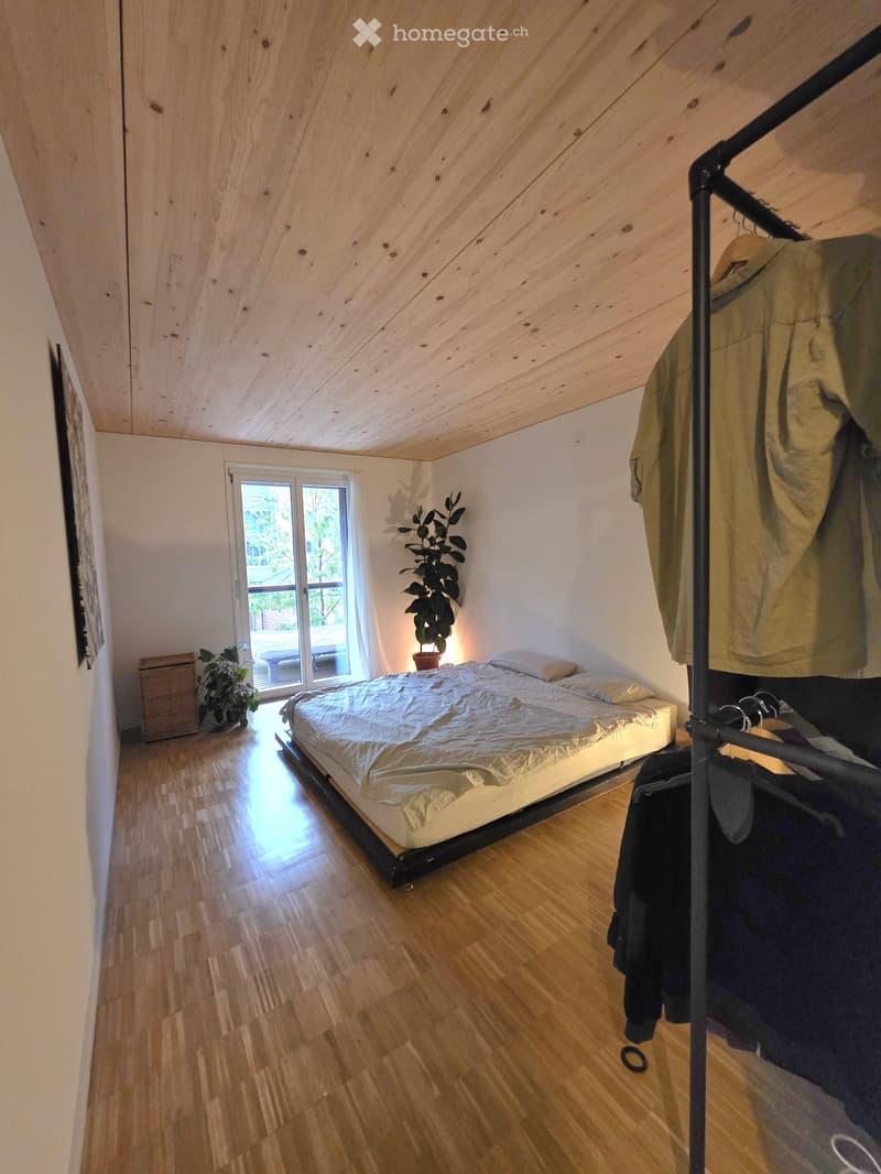 Cozy bedroom with wooden floors, slanted wooden ceiling, bed with white bedding, clothing rack, and potted plants near the window.