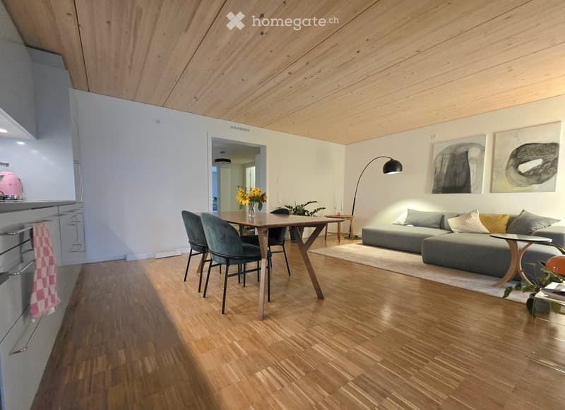 Open-plan living and dining area with wooden flooring, grey sectional sofa, wooden coffee table, dining table with chairs, floor lamp, and artwork on the walls.
