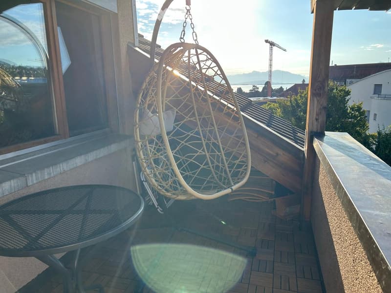Balcony with a hanging wicker chair and a small round table, overlooking a scenic view of mountains and a body of water.