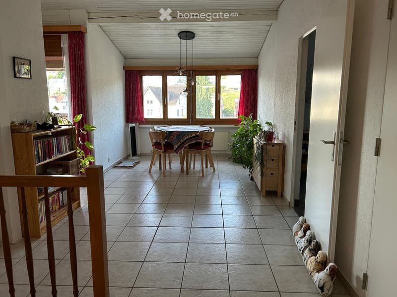 Bright dining area with tiled floor, large windows, wooden table, chairs, bookshelf, fan, and plants.