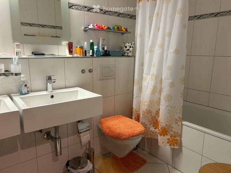 Bathroom with a sink, toilet, shower curtain, and various toiletries on the counter. Features a tiled wall and a small wooden stool near the toilet.