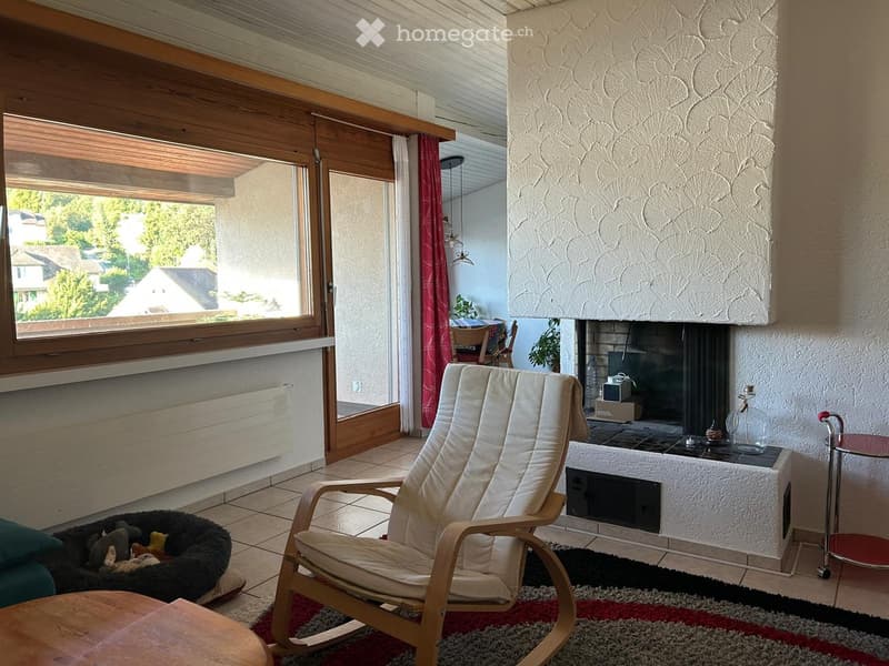 Cozy living room with a fireplace, large window with red curtains, a white armchair, a dog bed, and a small table.