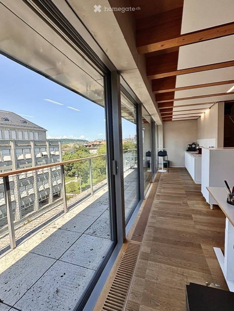 Spacious balcony with wooden flooring, large glass windows, and a view of the city and surrounding greenery.