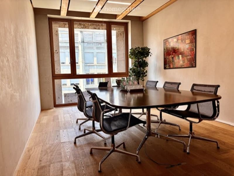Conference room with a large wooden table, several black office chairs, a potted plant, and a colorful abstract painting on the wall. The room has large windows with blinds and a polished wooden floor.