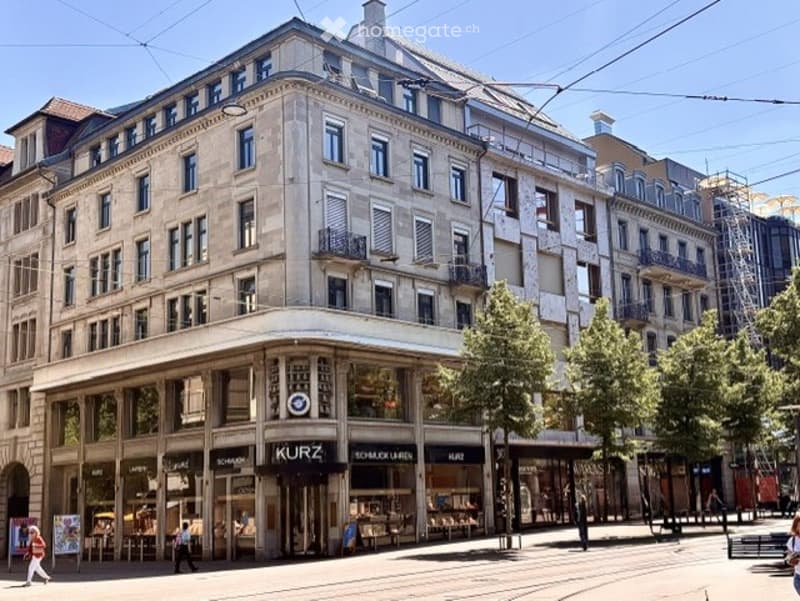 A multi-story urban building with classic architecture, large windows, and a storefront labeled "KURZ" at the ground level. The building features ornate detailing and is situated on a busy street with pedestrians and trees.