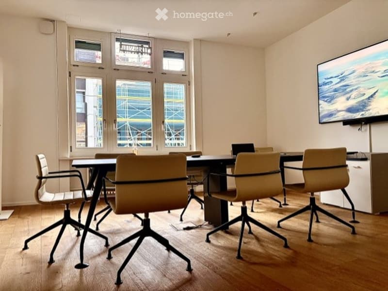 Modern office space with a large conference table, ergonomic chairs, a laptop, and a large screen displaying an image.