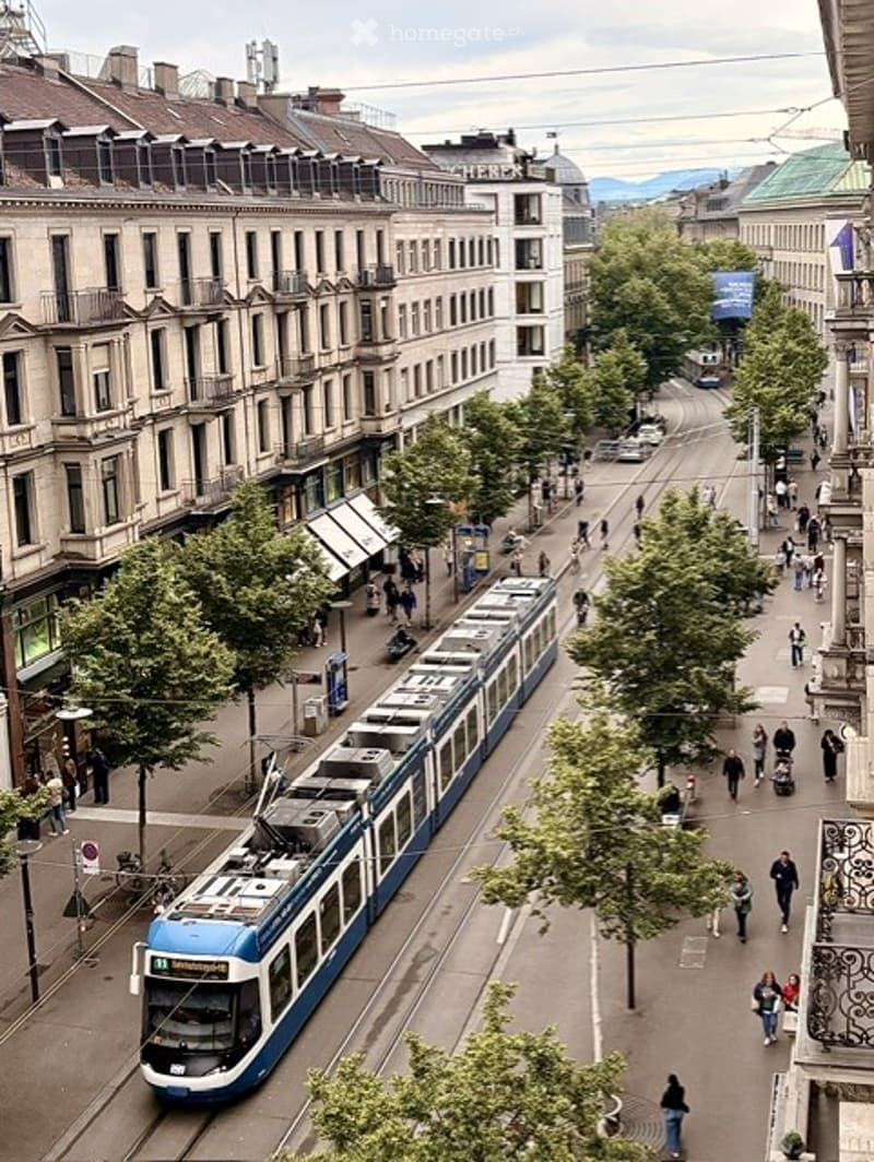 A bustling urban street lined with multi-story buildings, featuring a modern tram moving along the tracks, pedestrians walking on the sidewalks, and trees providing shade.