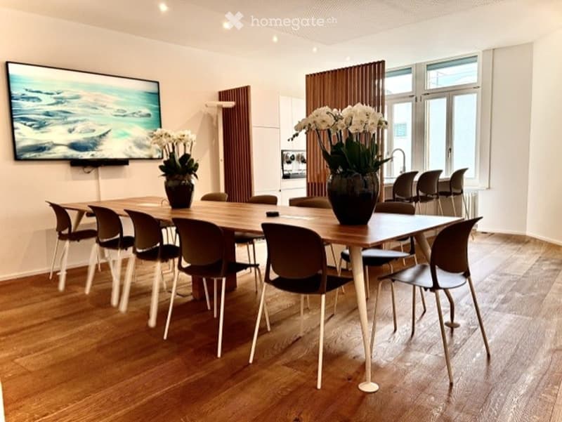 Modern dining room with a large wooden dining table surrounded by black chairs, two vases with white flowers, and a television mounted on the wall.