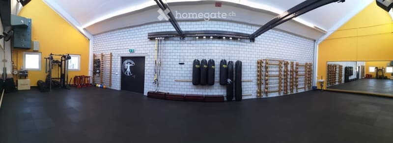 A spacious gym room with a black rubber floor, white tiled walls, and yellow accent walls. The room features a large mirror, various weights and exercise equipment, and several punching bags hanging from the wall.