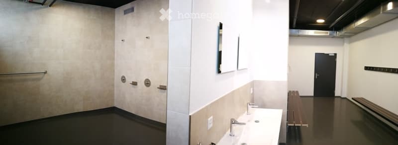 Empty locker room with white walls, dark flooring, and a row of sinks. There are benches and a closed door visible.