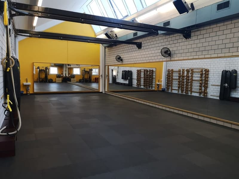 A spacious gym room with dark flooring, large mirrors, weight racks, and punching bags. The room features exposed beams and bright lighting.