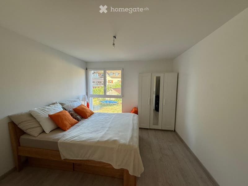 A well-lit bedroom with a wooden bed, white bedding, orange pillows, a wardrobe, and a small lamp on a bedside table. The room has light-colored walls and wooden flooring.