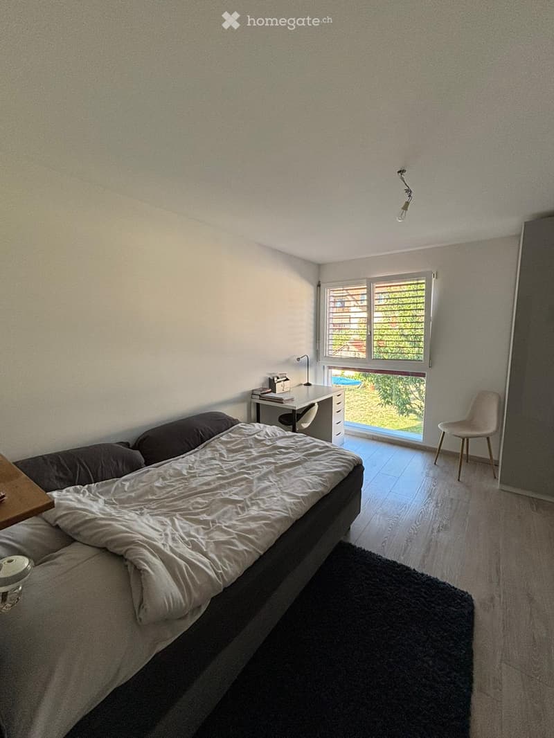 A modern bedroom with a bed, desk, chair, and a large window with blinds. The room features a dark rug and a wooden side table.