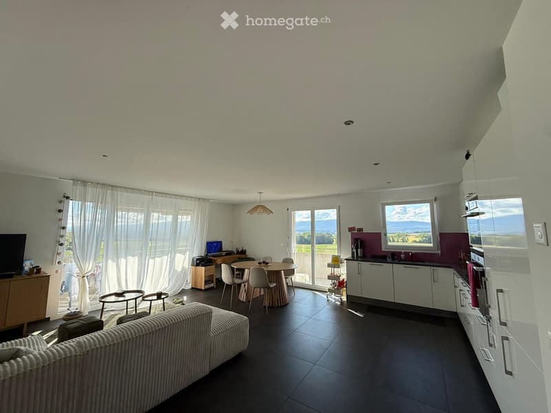 Open-plan living and dining area with a sofa, coffee table, dining table, chairs, and a modern kitchen with cabinets, refrigerator, and oven. Large windows offer a scenic view.