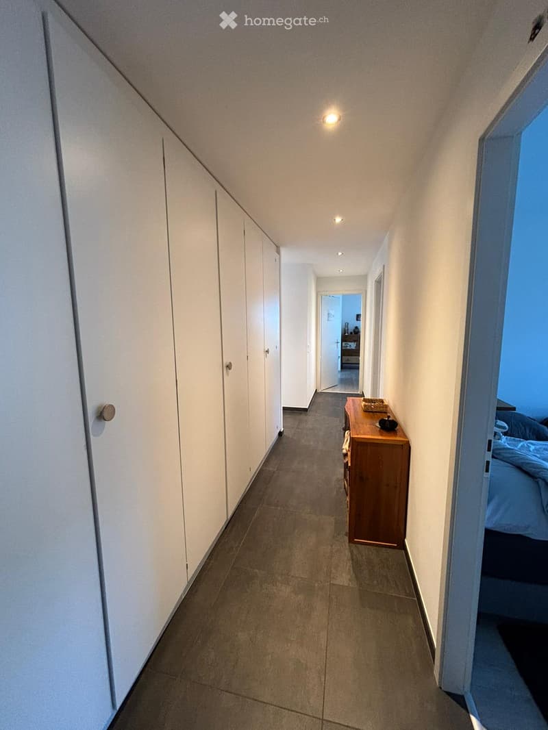 A modern hallway with white walls, grey tiled floor, built-in white cabinets on one side, and a small wooden console table with decorative items on the other side. The hallway leads to a bedroom with a bed visible at the end.