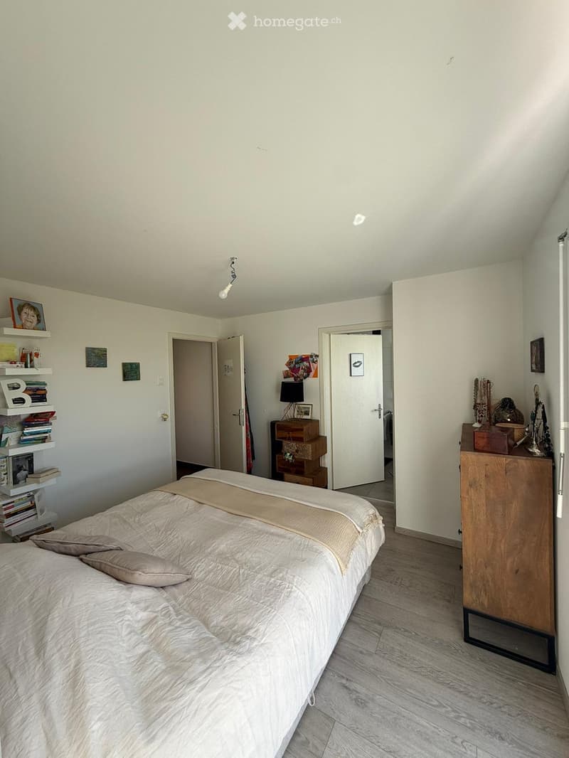A modern bedroom with a large bed, nightstand, shelf with books and decorative items, and a lamp on a small table.