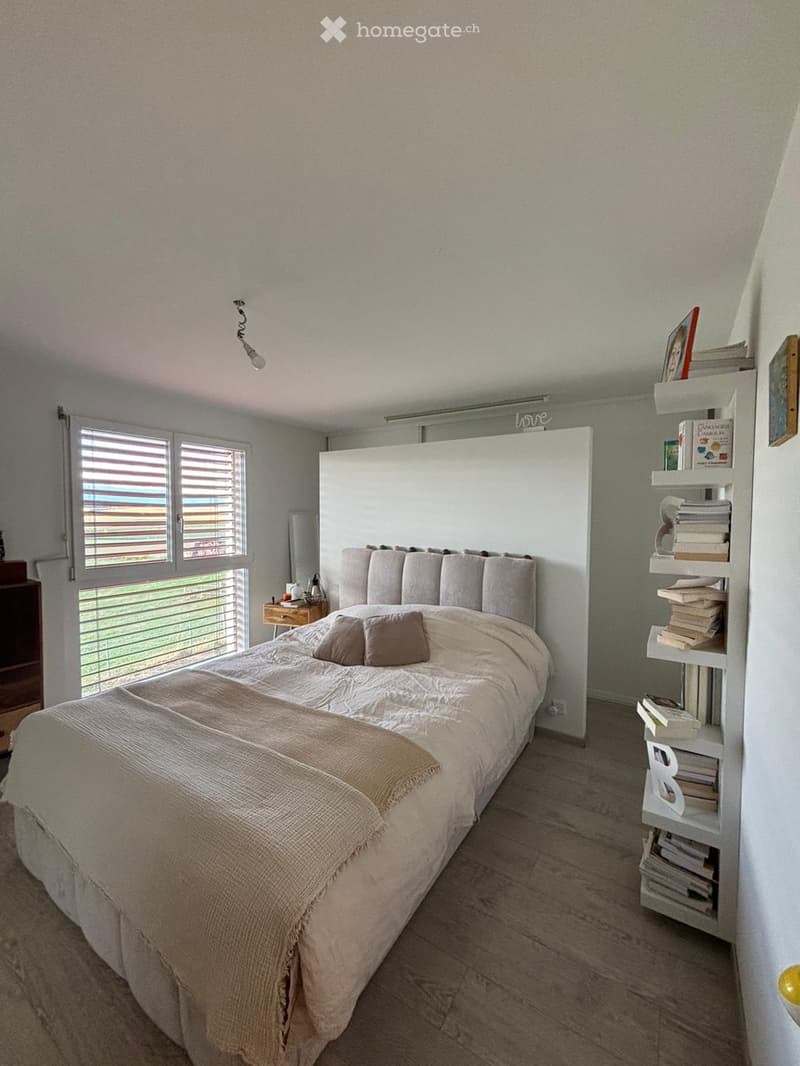 A modern bedroom with a large bed, nightstand, and bookshelf filled with books. The room features wooden flooring and large windows with blinds.