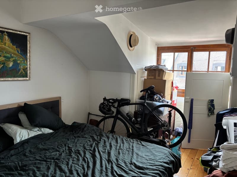 A cozy bedroom with a slanted ceiling, featuring a bed with dark bedding, a bicycle leaning against the wall, a painting on the wall, and a hat hanging on the wall. There is also a refrigerator and some clothing items visible.