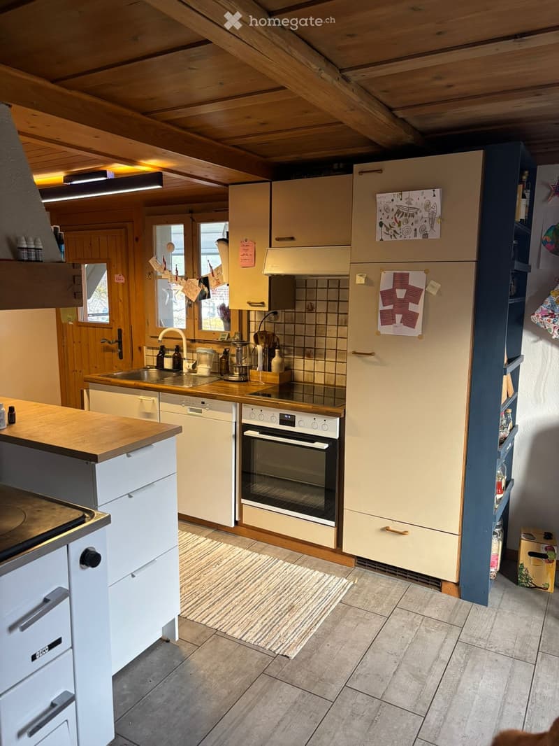 Bright kitchen with wooden ceiling beams, light-colored cabinets, and modern appliances including an oven, refrigerator, and dishwasher. Wooden countertops and tiled floor.
