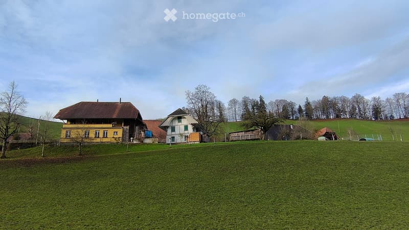 A two-story house with a yellow exterior and a brown roof, situated on a grassy field with trees and hills in the background.