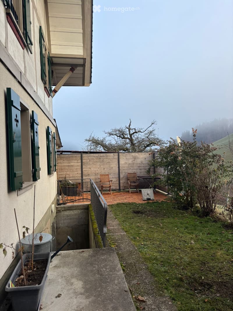 Outdoor garden area with a wooden fence, small patio with chairs and a table, grassy lawn, and a view of a misty landscape.