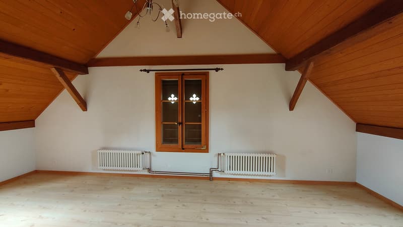 Empty attic room with wooden slanted ceilings, wooden floor, large window with double glass panels, and two radiators on either side of the window.