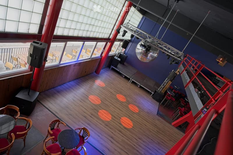 A spacious room with wooden flooring, large windows, and a stage area with a disco ball and speakers. Tables and chairs are arranged for seating, and the room features a balcony area visible through the windows.