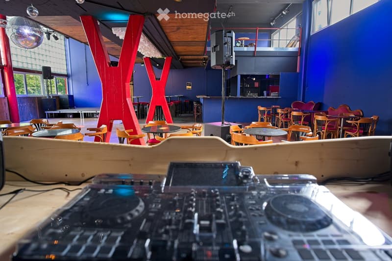 A modern, spacious room with wooden floors, blue walls, and large windows. The room features multiple tables and chairs arranged for seating, a bar area with stools, and a DJ turntable on a wooden platform. Red structural elements and a disco ball add a v