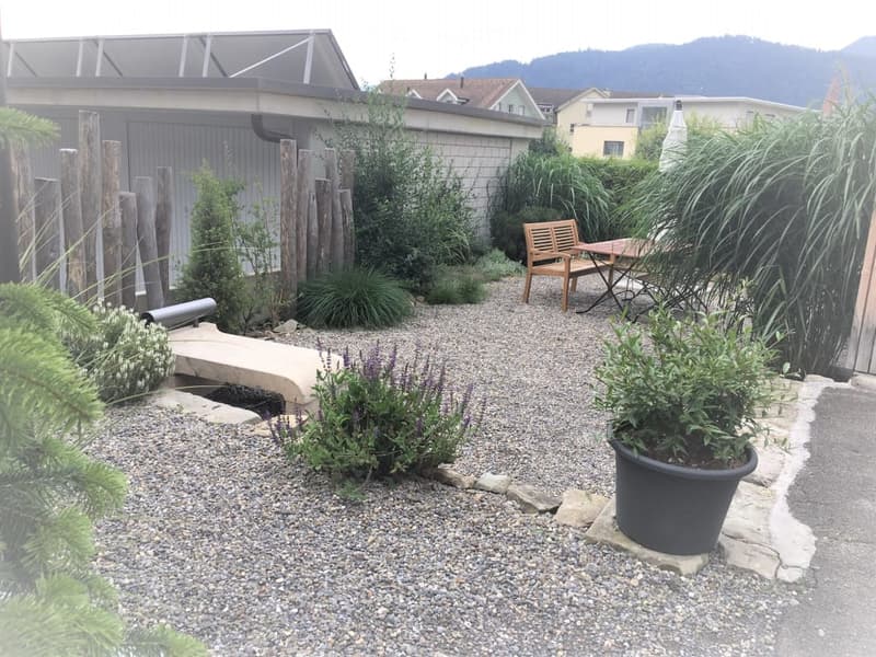 Garden area with gravel pathway, wooden bench, small table and chairs, various plants and shrubs, and a modern building in the background.