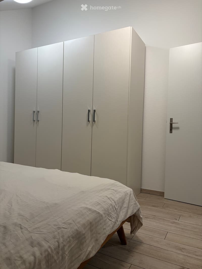 A modern bedroom with a wooden floor, a neatly made bed, and a large white wardrobe with mirrored doors.