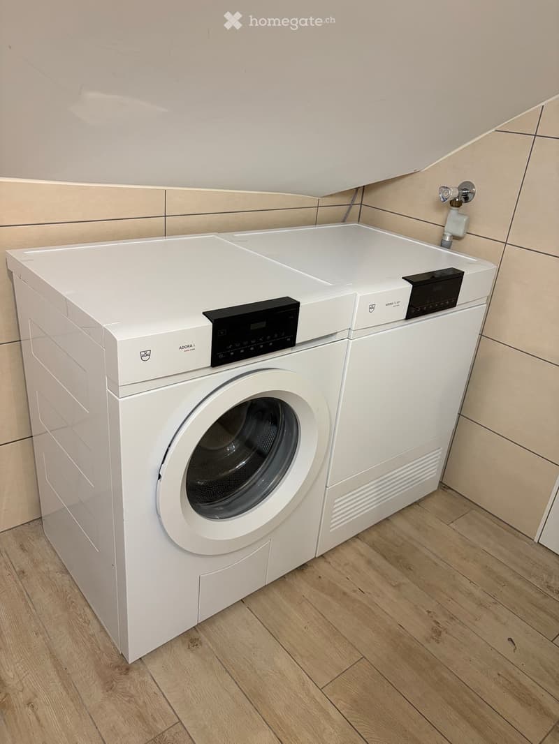 White washing machine with digital control panel, placed on wooden floor in a tiled bathroom.