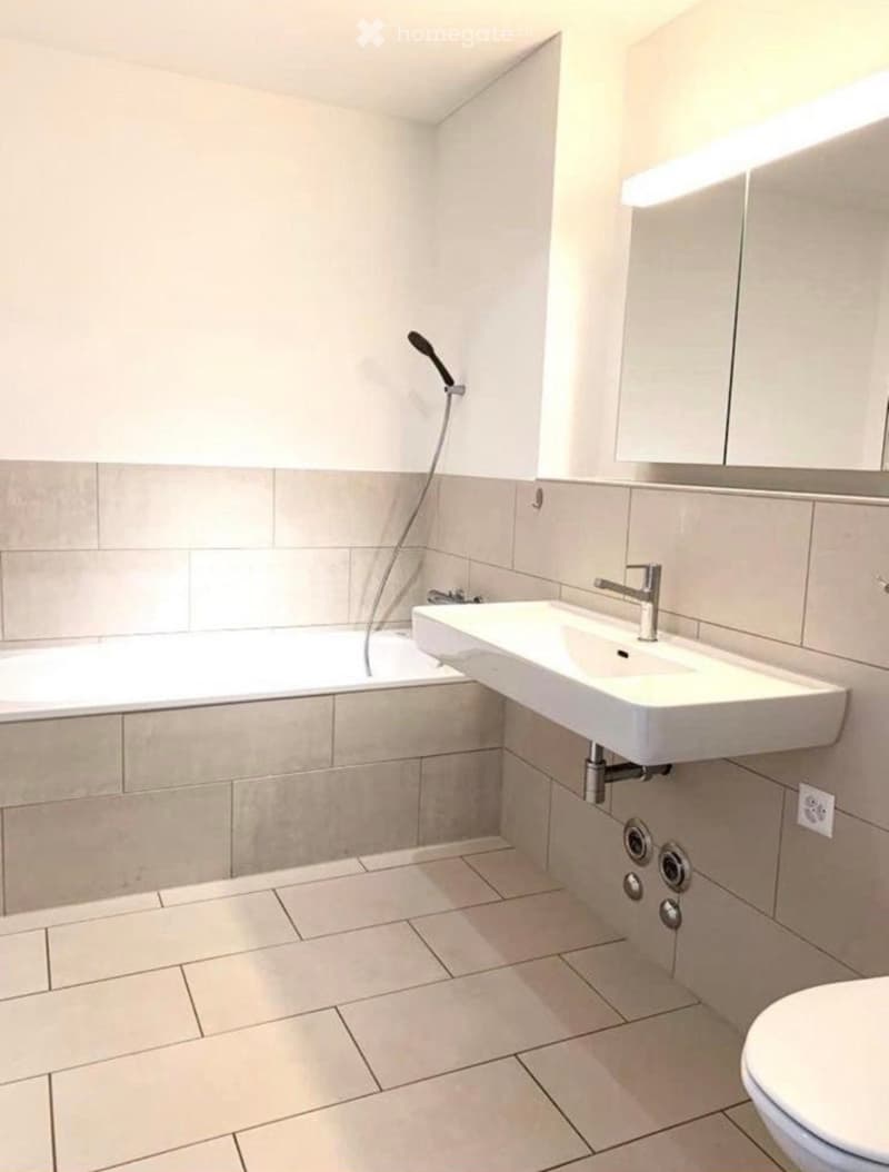 Modern bathroom with a bathtub, sink, and toilet. Features tiled walls and floor, a handheld showerhead, and a modern faucet.